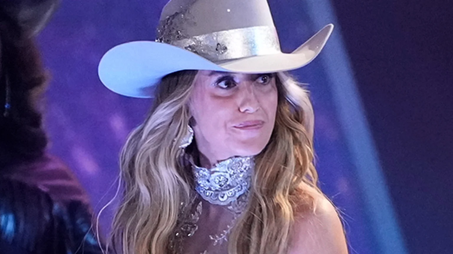A woman with long blonde hair wears a grey and light blue cowboy hat decorated with silver and a sparkly silver choker, looking to her right in a blurry purple and blue background.