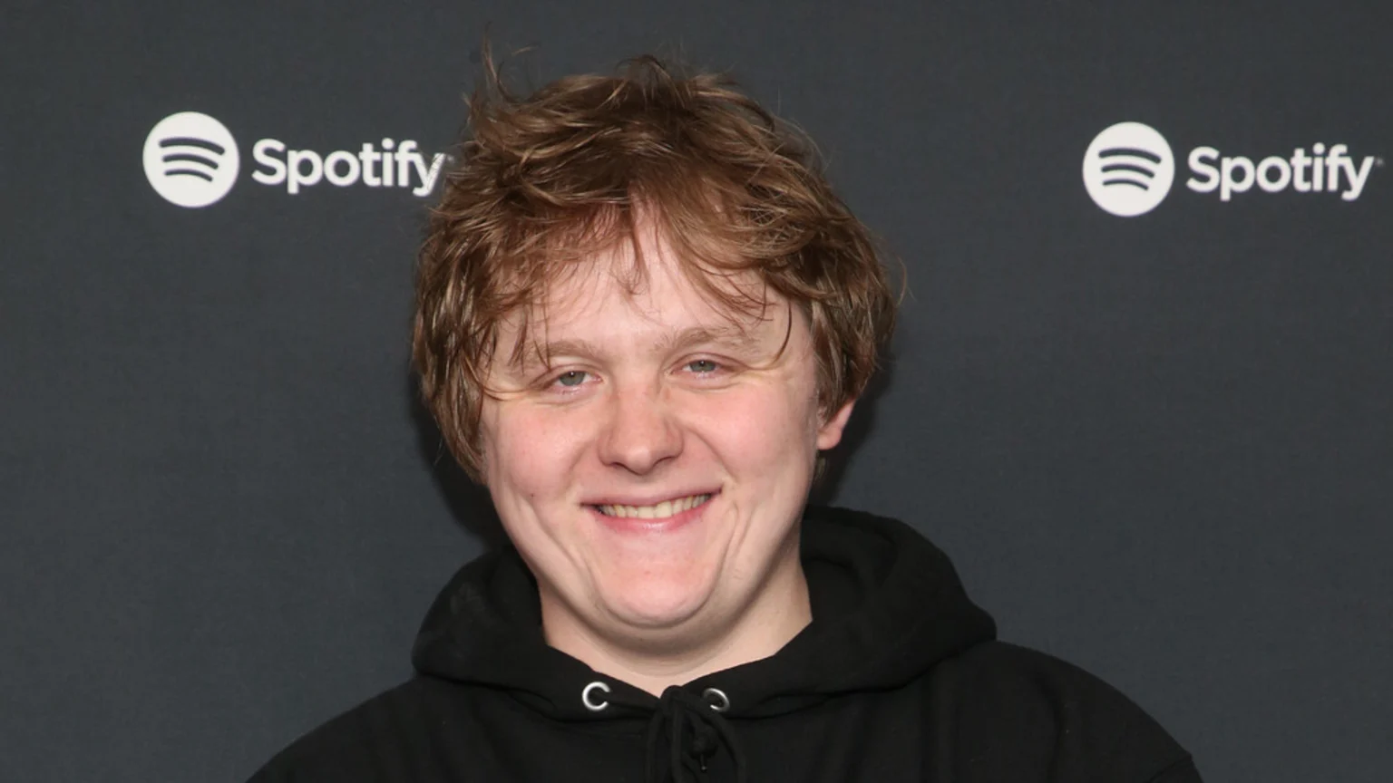 A smiling man with messy, light brown hair and a black hoodie stands in front of a dark grey background with two white Spotify logos.