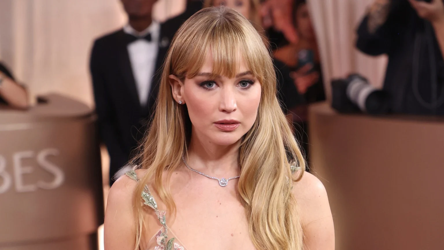 Jennifer Lawrence, with blonde hair and bangs, wears a light embroidered dress and a diamond necklace, looking at the camera with a neutral expression at an event.