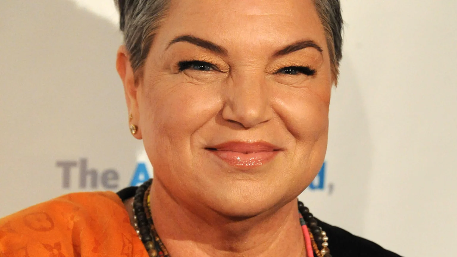 A close-up shot of a smiling woman with short gray hair, golden eyeshadow, and a subtle earring, wearing an orange top and beaded necklaces.