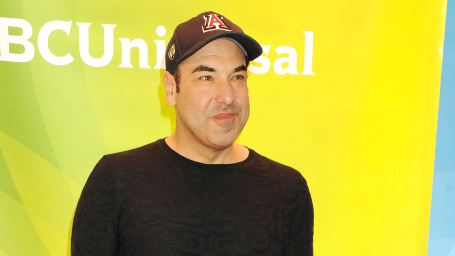 A man in a black long-sleeved shirt and a dark baseball cap with a red 'A' logo stands against a yellow-green background with "NBCUniversal" text.