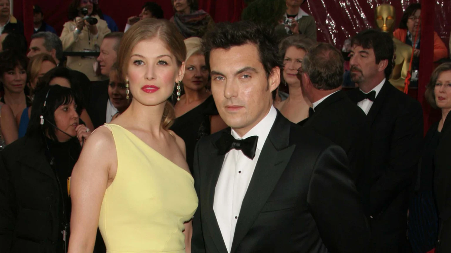 Rosamund Pike in a yellow dress and Joe Wright in a tuxedo pose on a red carpet with other people in the background, including a golden Oscar statue.