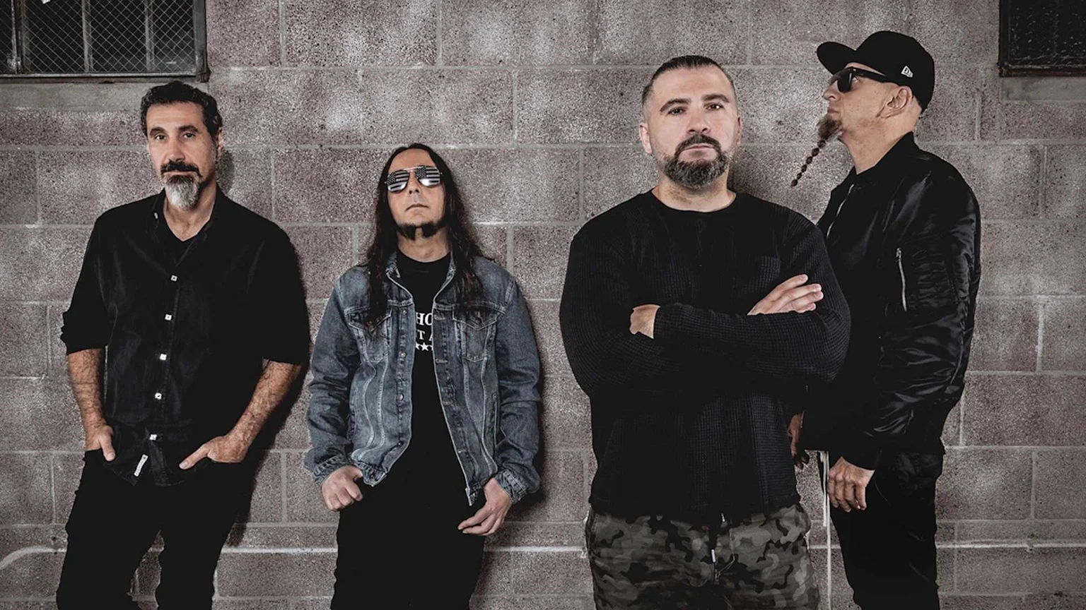 Four men, identified as System Of A Down members, stand against a brick wall, wearing casual dark clothing and various facial hair styles.