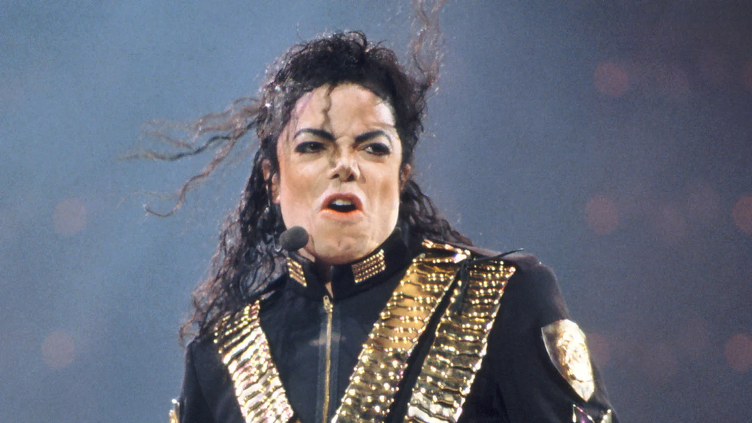 Michael Jackson performs on stage, wearing a black military-style jacket with gold embellishments, a microphone near his mouth, and an intense, expressive look.