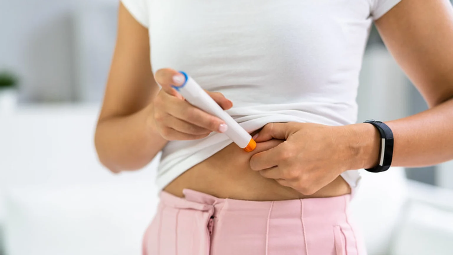 A person in a white shirt and pink pants self-injects into their abdomen with a white pen.