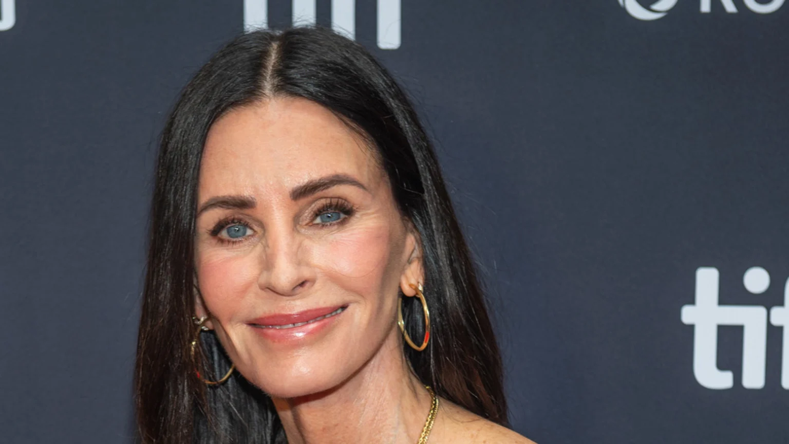 Courteney Cox with a gentle smile, long dark hair parted in the middle, blue eyes, makeup, gold hoop earrings, and a gold necklace against a dark background.