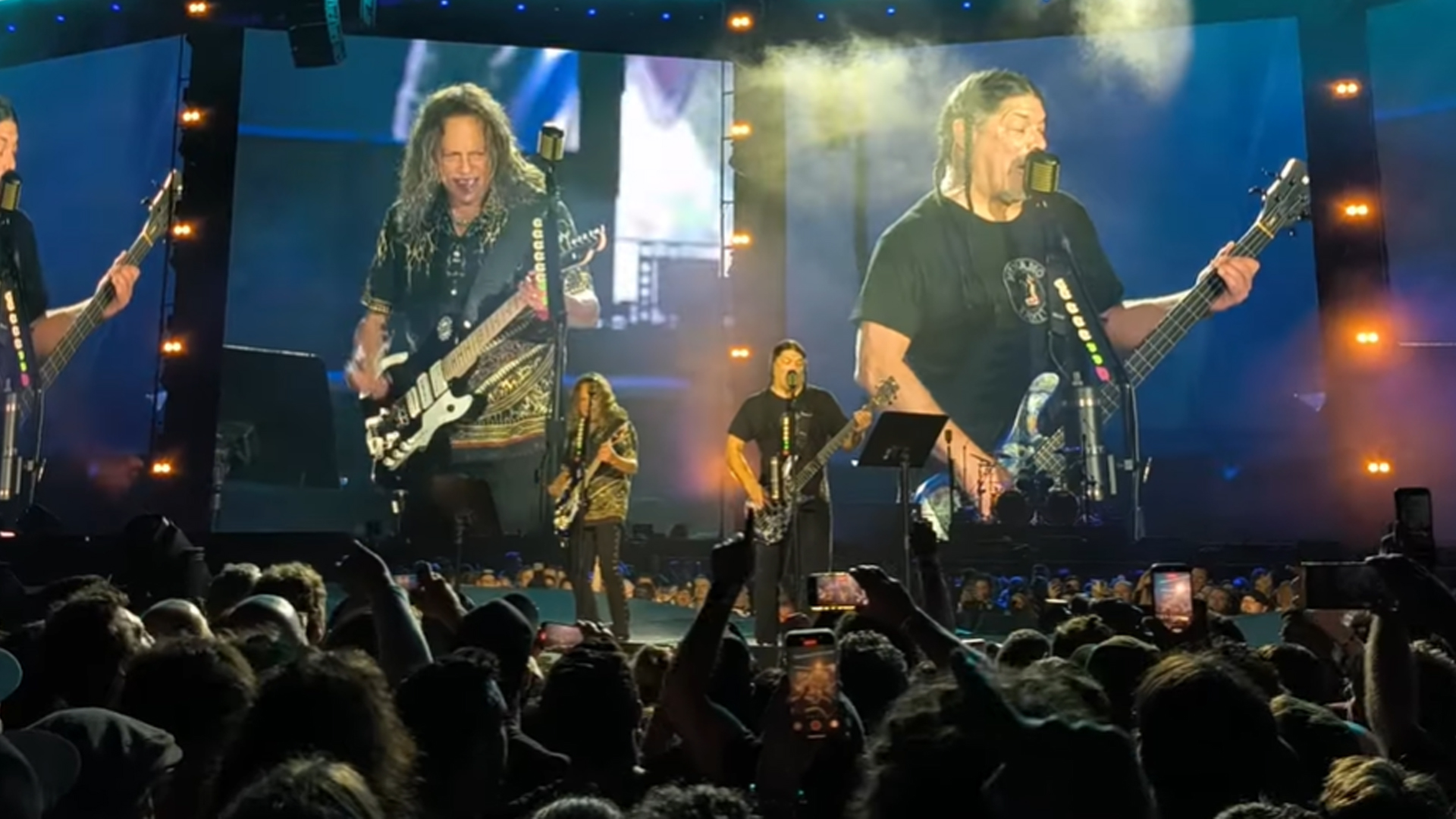 Kirk Hammett and Rob Trujillo covering The Chats' song Smoko at Brisbane Show. They are standing on stage playing the guitar with a big crowd. 