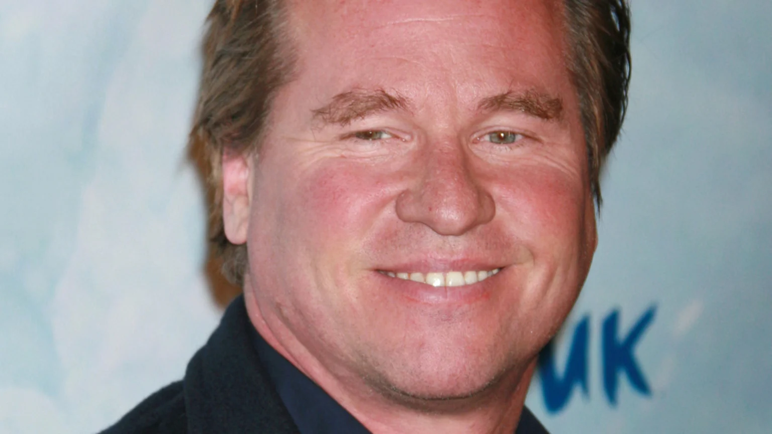 A man with reddish-blond hair and flushed skin smiles, wearing a dark jacket against a light blue background with faint blue letters "MK".