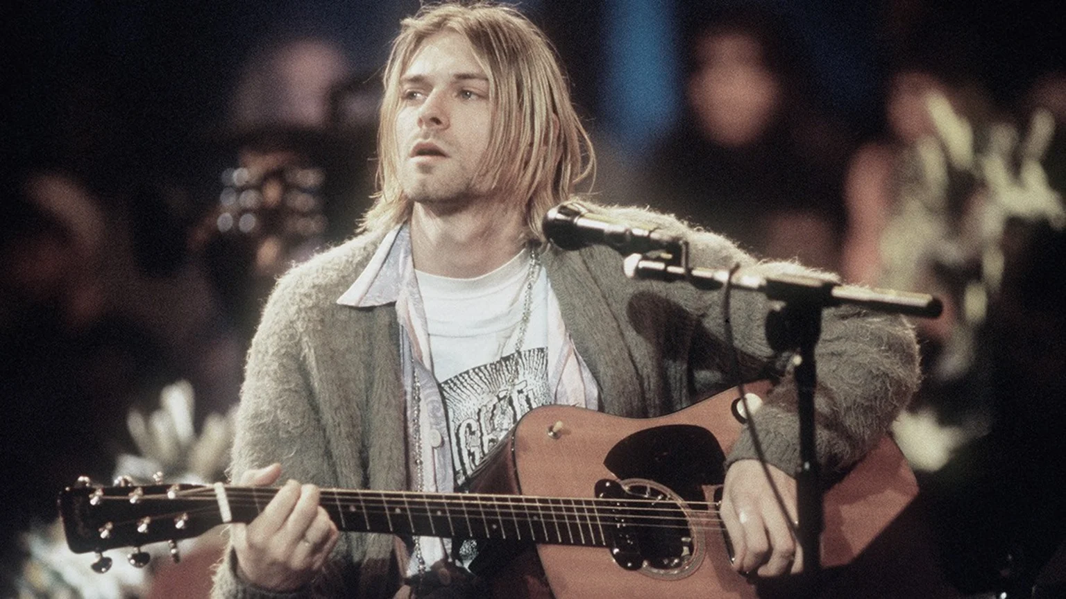Medium shot of Kurt Cobain in his twenties on a dimly lit indoor stage, playing a brown acoustic guitar and singing into a dark microphone. He wears a green fuzzy cardigan, layered shirts, a silver beaded necklace, and a plain silver ring.