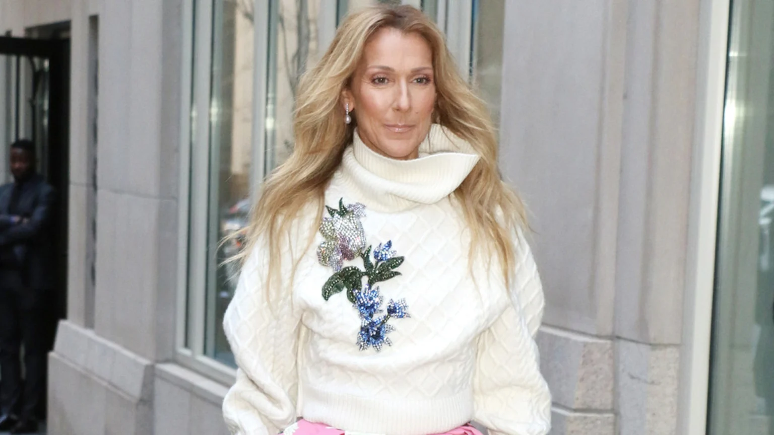 Celine Dion, with long blonde hair and pearl earrings, wears a cream cable-knit turtleneck sweater with a sparkling floral embellishment, standing outdoors.