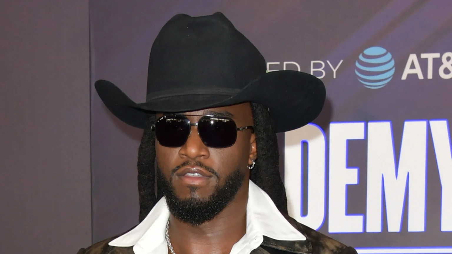 A man with dark skin wears a black cowboy hat, dark sunglasses, and has a beard and dreadlocks, looking forward against a blurry purple background with white text and a blue AT&T logo.