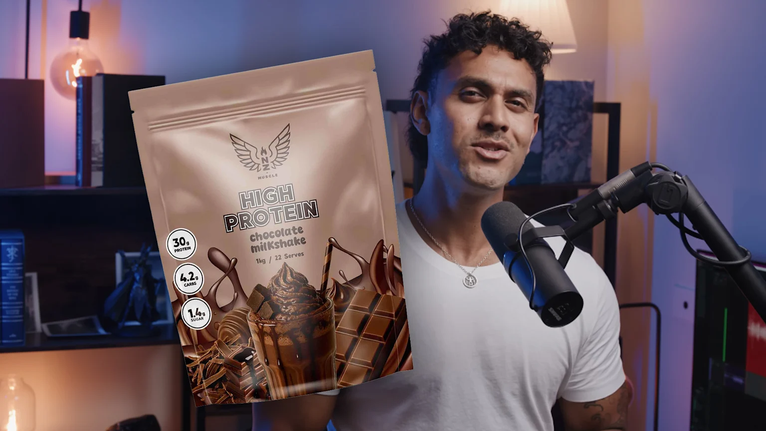 A man in a white t-shirt speaks into a microphone, while a superimposed NZ Muscle High Protein chocolate milkshake pouch, displaying nutritional info and chocolate imagery, covers part of the frame.