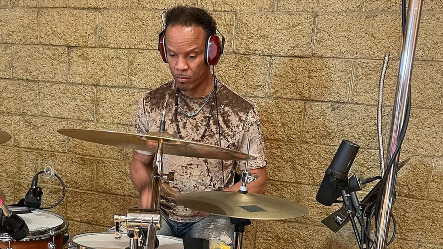 A male drummer with headphones and a brown textured shirt plays drums, with cymbals and a microphone visible, against a beige brick wall.