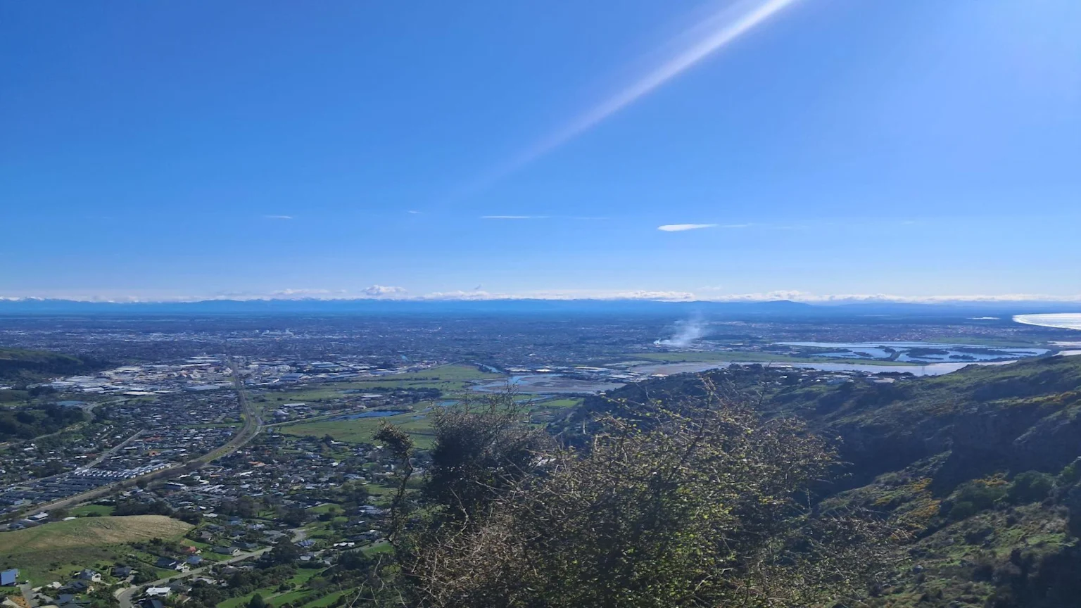 A panoramic view from a hillside overlooks a large city, river, and estuary under a bright blue sky with snow-capped mountains in the distance and a plume of smoke rising from an area on the right.
