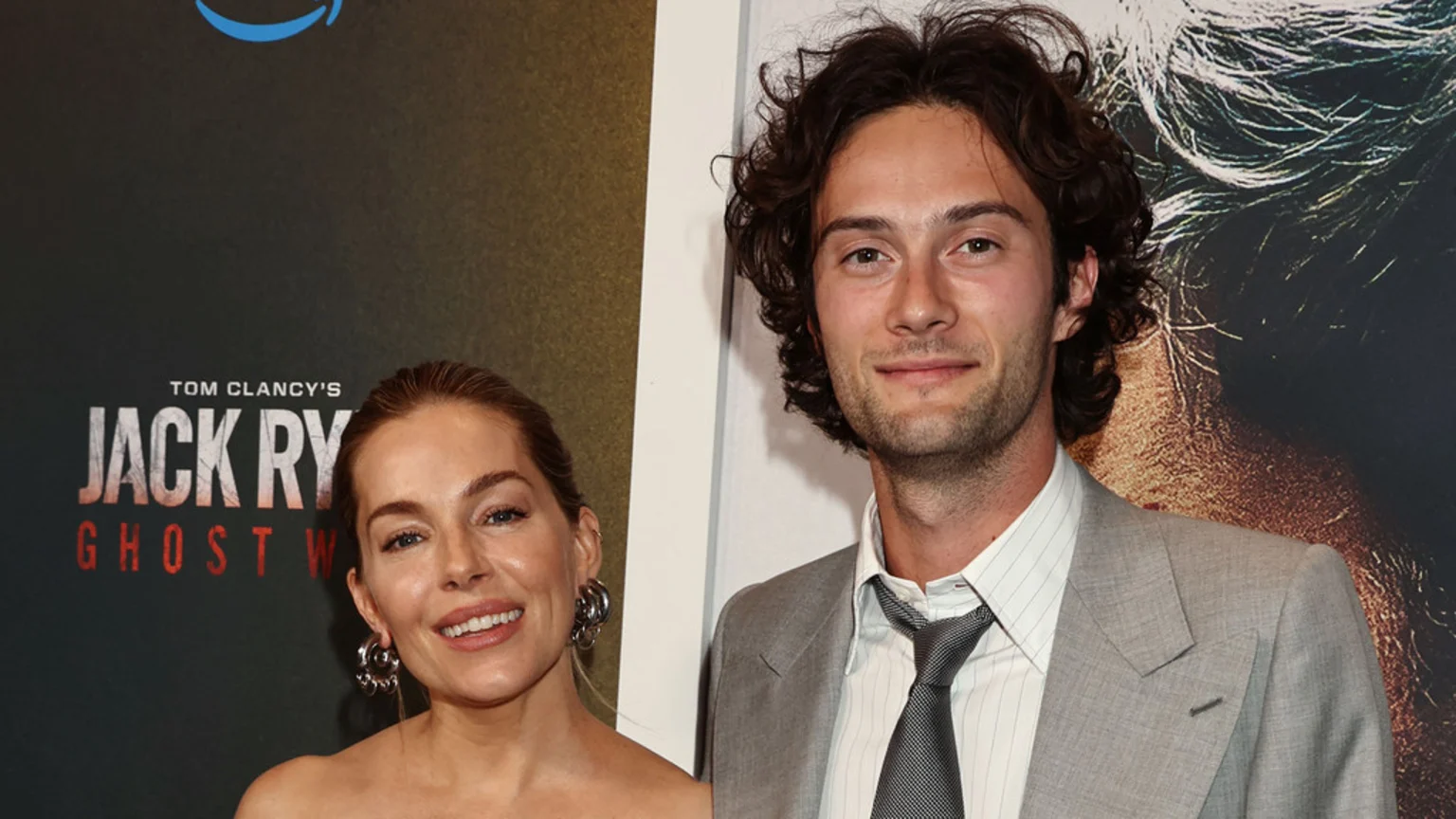 Sienna Miller and Oli Green smile, standing side-by-side in front of a "Tom Clancy's Jack Ryan" poster with the Amazon Prime Video logo.