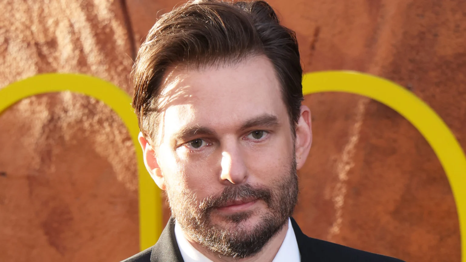 A man with dark hair, a beard, and fair skin under direct sunlight looks forward, wearing a dark suit jacket and white shirt against a blurry orange background with yellow curves.