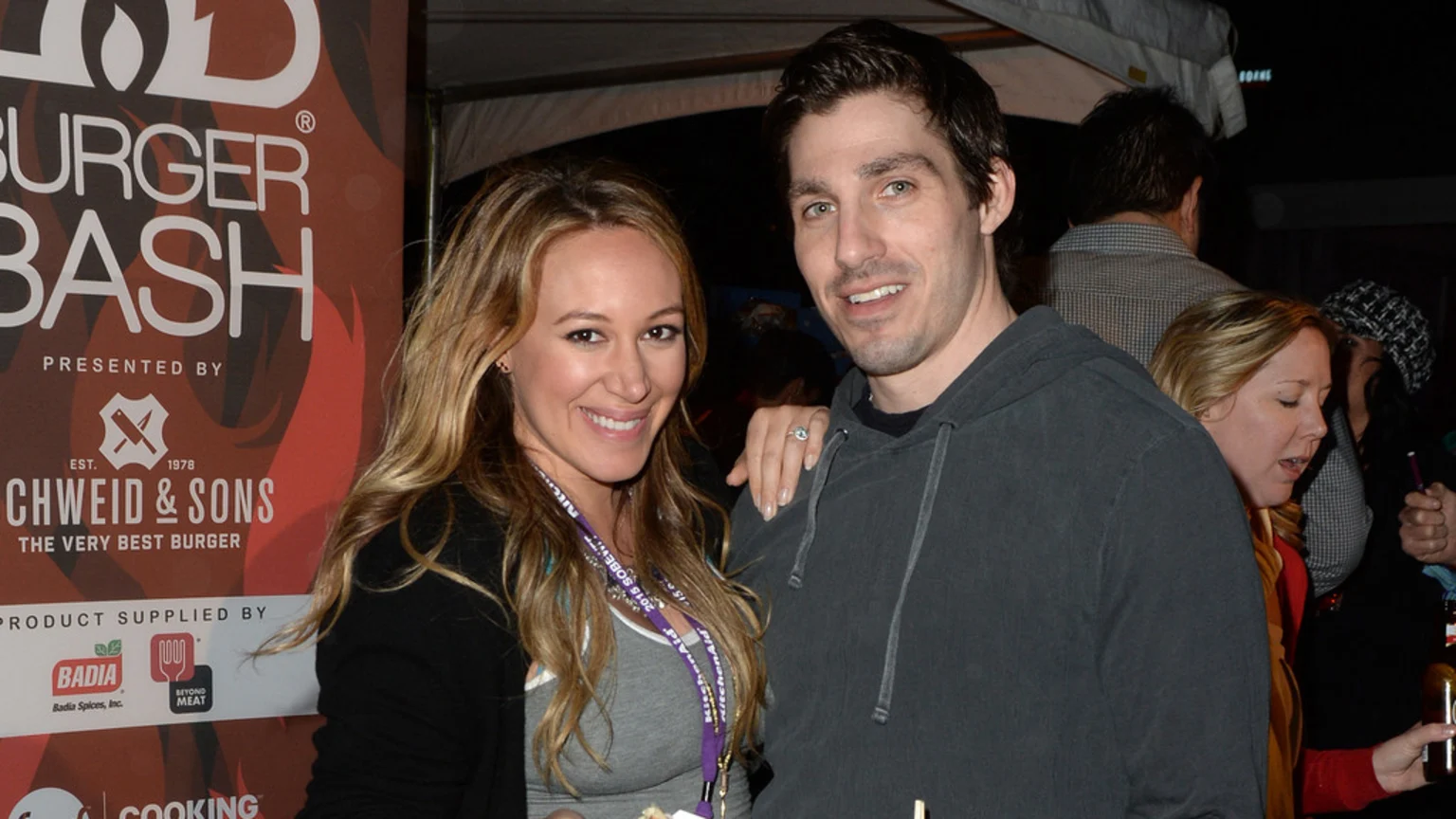 A smiling woman with long blonde hair and a ring on her left hand stands next to a smiling man in a dark hoodie, with a "Burger Bash" banner behind them.