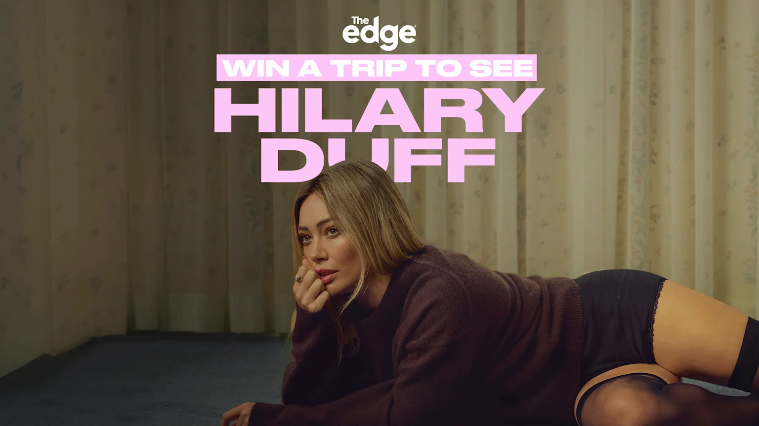 Hilary Duff lies on a blue carpet in a brown sweater and black lingerie, looking to the side, with a floral wall behind her and text announcing 'WIN A TRIP TO SEE HILARY DUFF'.