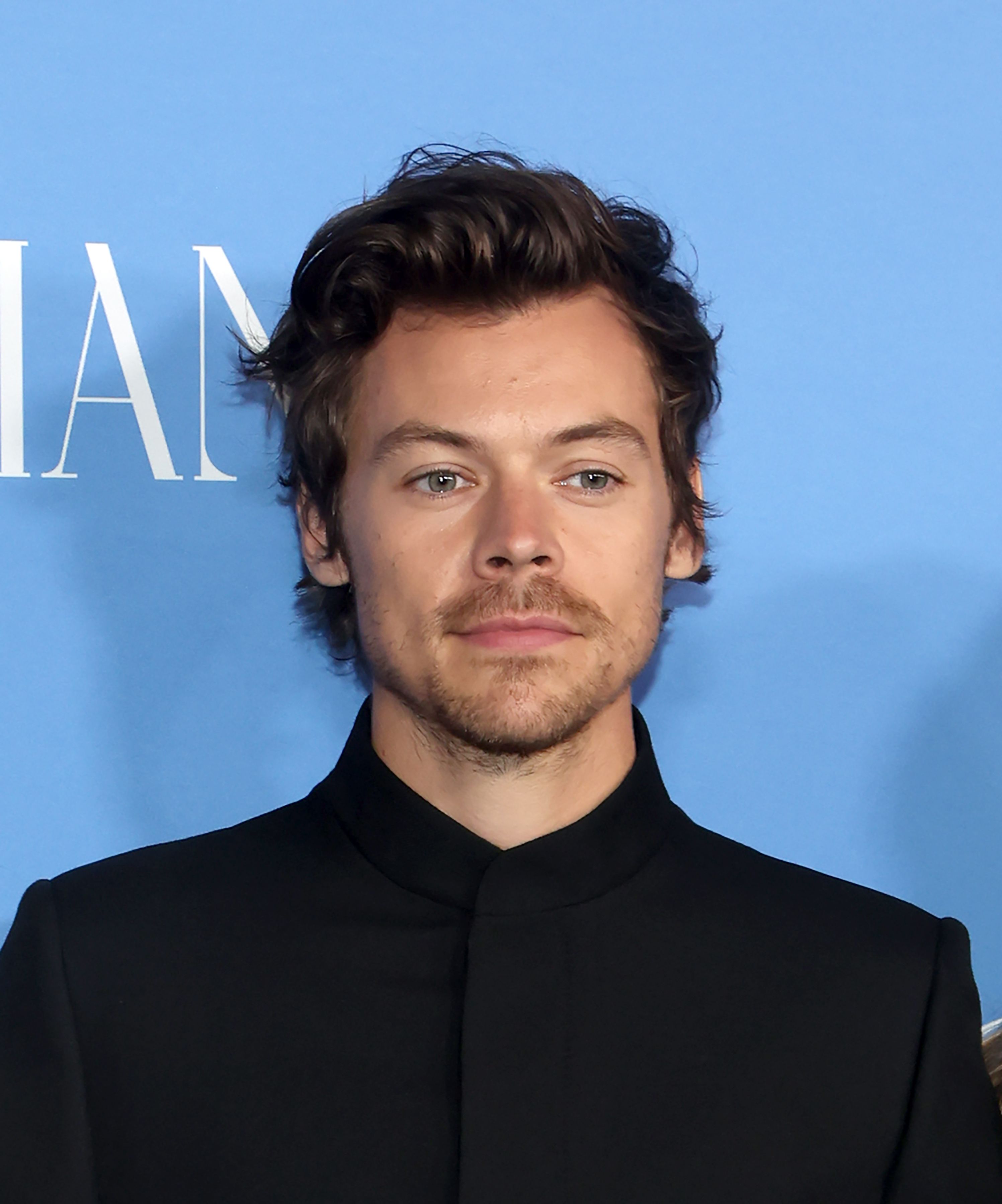 A close-up headshot of Harry Styles with brown wavy hair, light eyes, and a light beard, wearing a black collared shirt, against a blue background with partial white text.