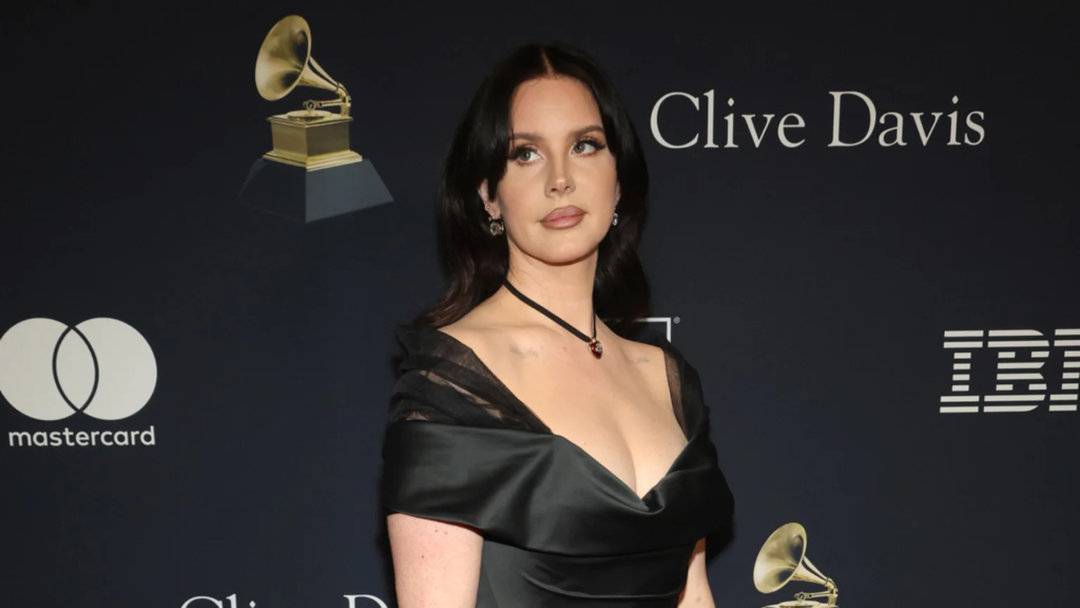 Lana Del Rey in a black off-the-shoulder dress looks to her right, standing against a dark backdrop featuring event and sponsor logos including a Grammy award, Mastercard, Clive Davis, and IBM.