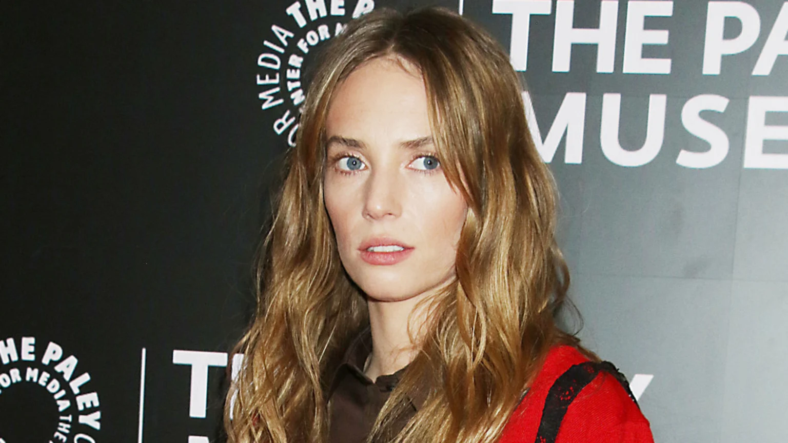 A woman with long wavy blonde hair and blue eyes, wearing a brown shirt and red jacket, stands in front of a dark background with "THE PALEY CENTER FOR MEDIA" and "THE PA MUSEUM" visible.