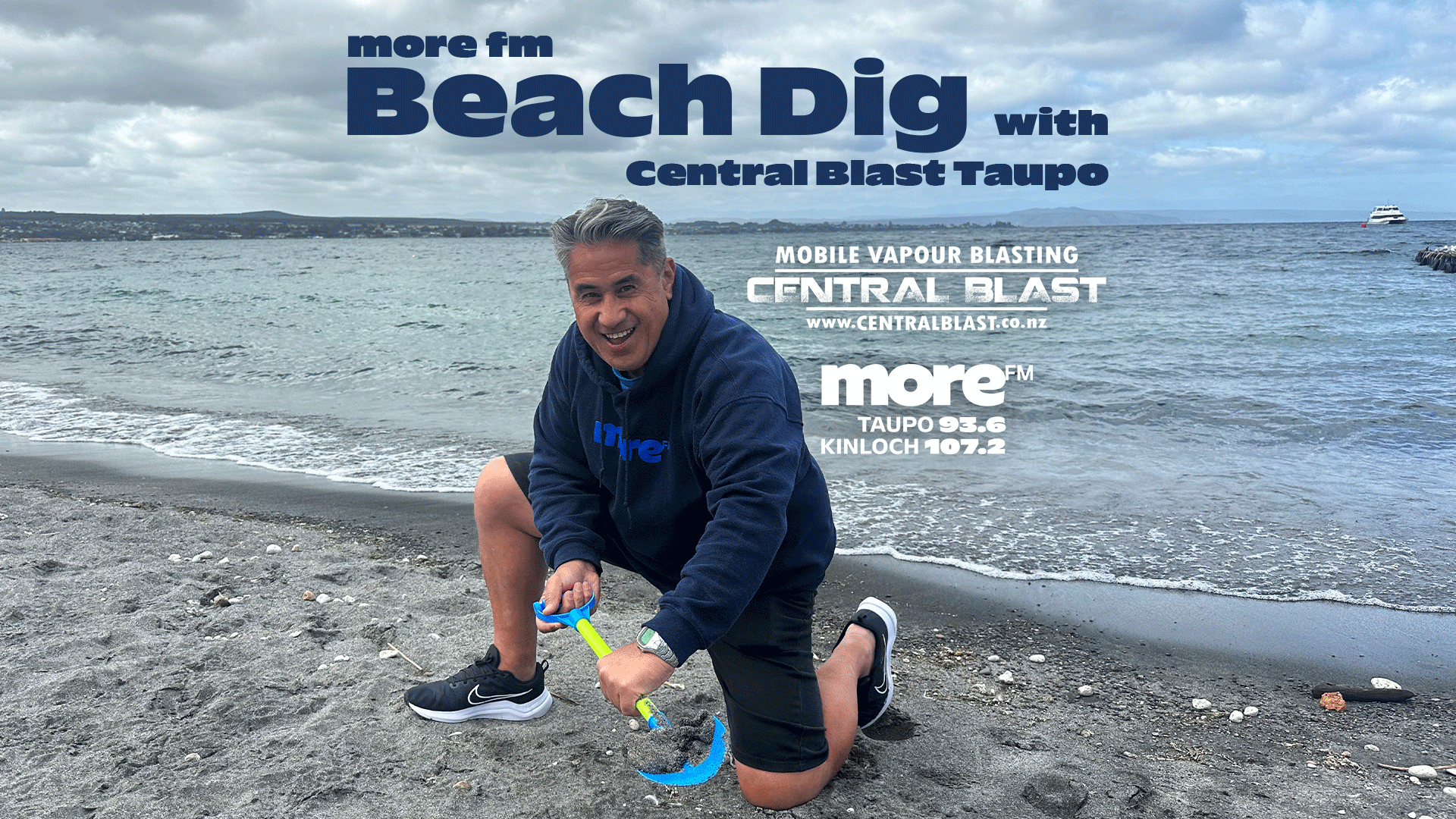 A smiling man in a more fm hoodie kneels on a dark sand beach with a small shovel, against a lake background, with "more fm Beach Dig with Central Blast Taupo" text overlay.