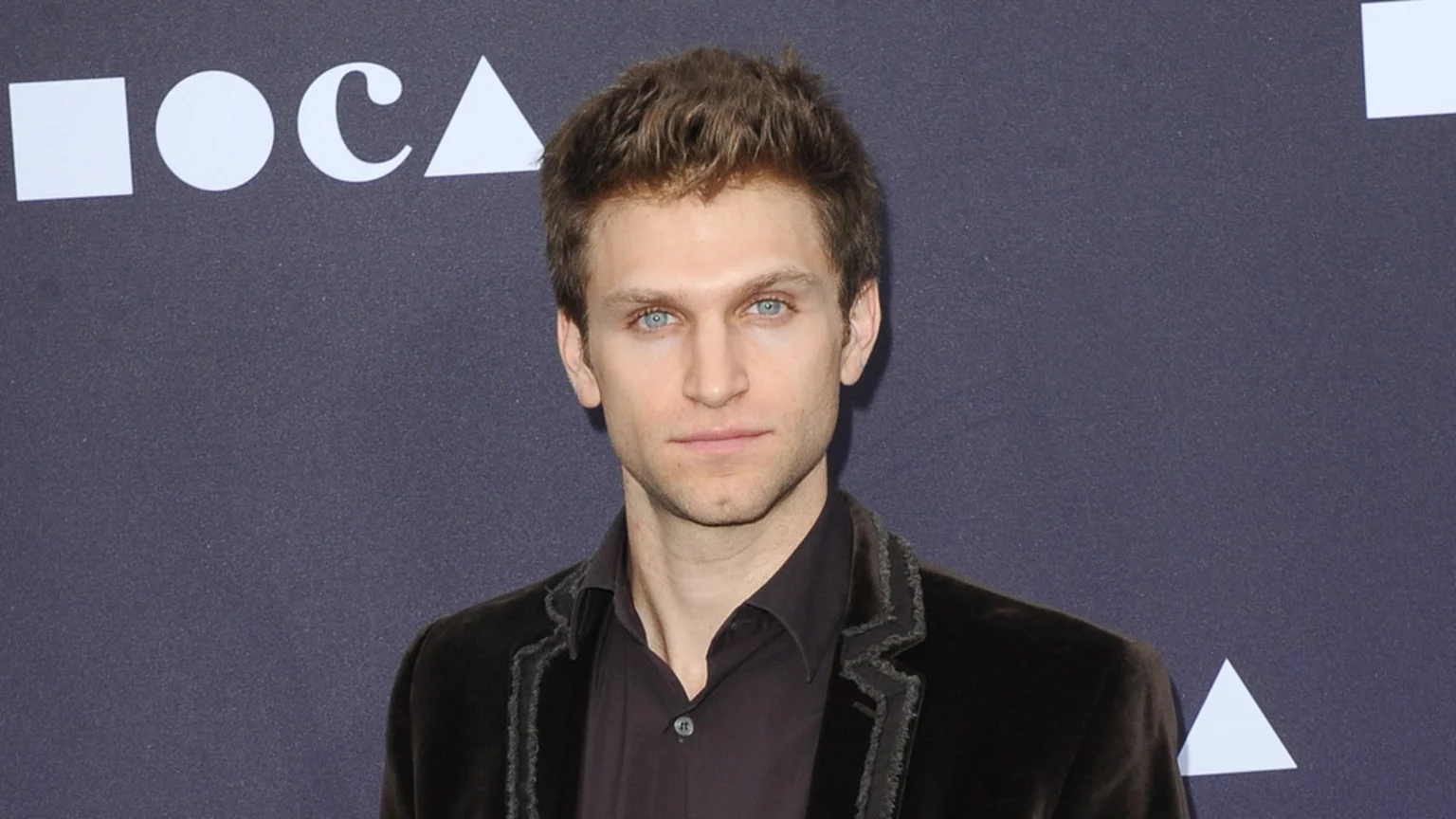 Keegan Allen, a man with light brown hair and blue eyes, looks directly at the camera, wearing a dark shirt and velvet jacket. A partial MOCA logo is visible in the top left background.