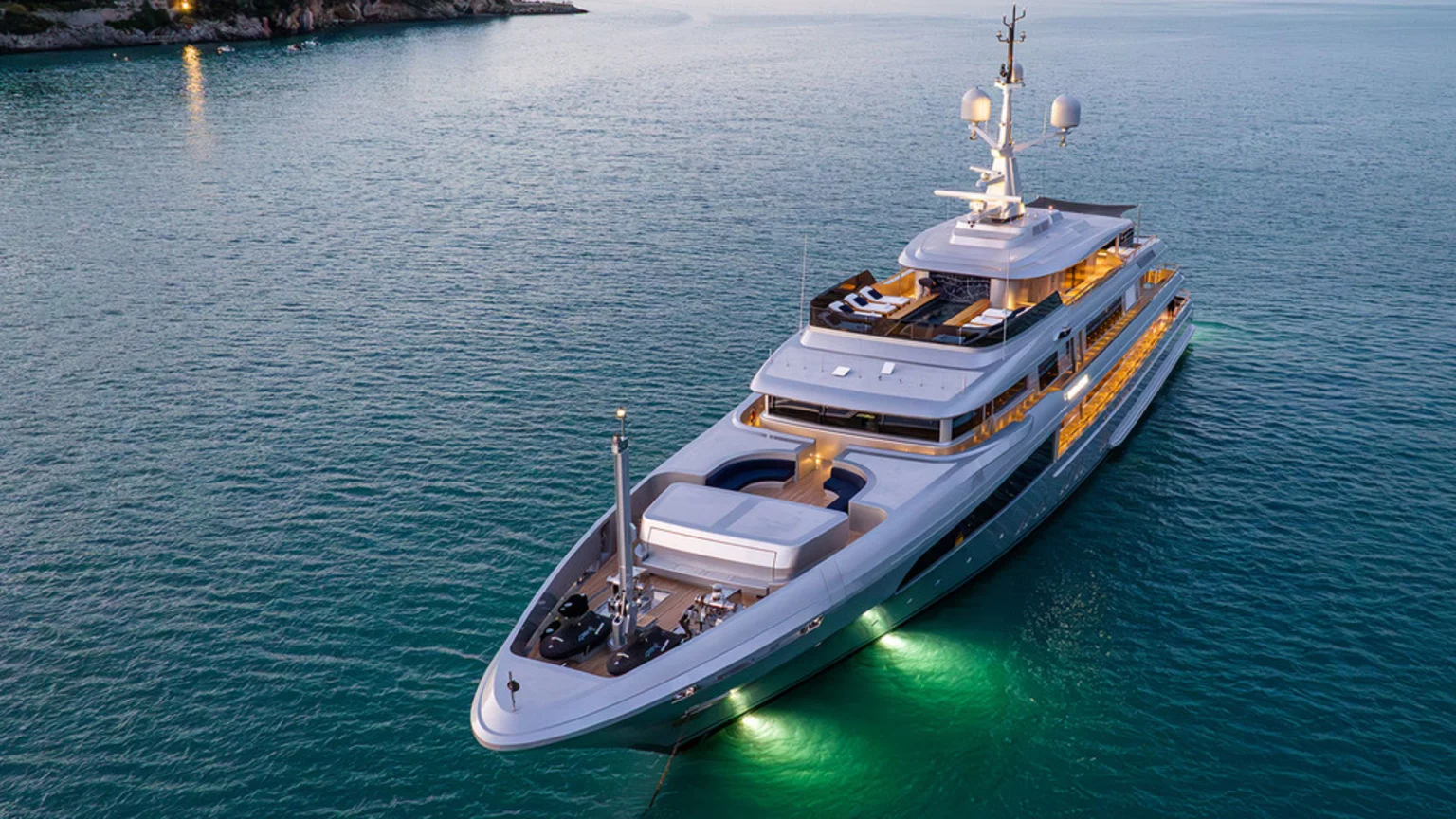 An aerial photo of a large modern multi-deck luxury yacht at anchor in a calm coastal bay during twilight. The yacht features warm interior and green underwater lights, multiple sun loungers on the top deck, a tall mast with communication equipment, and several watercraft stowed on a wooden bow deck. A rocky coastline with illuminated structures is visible in the distant background.