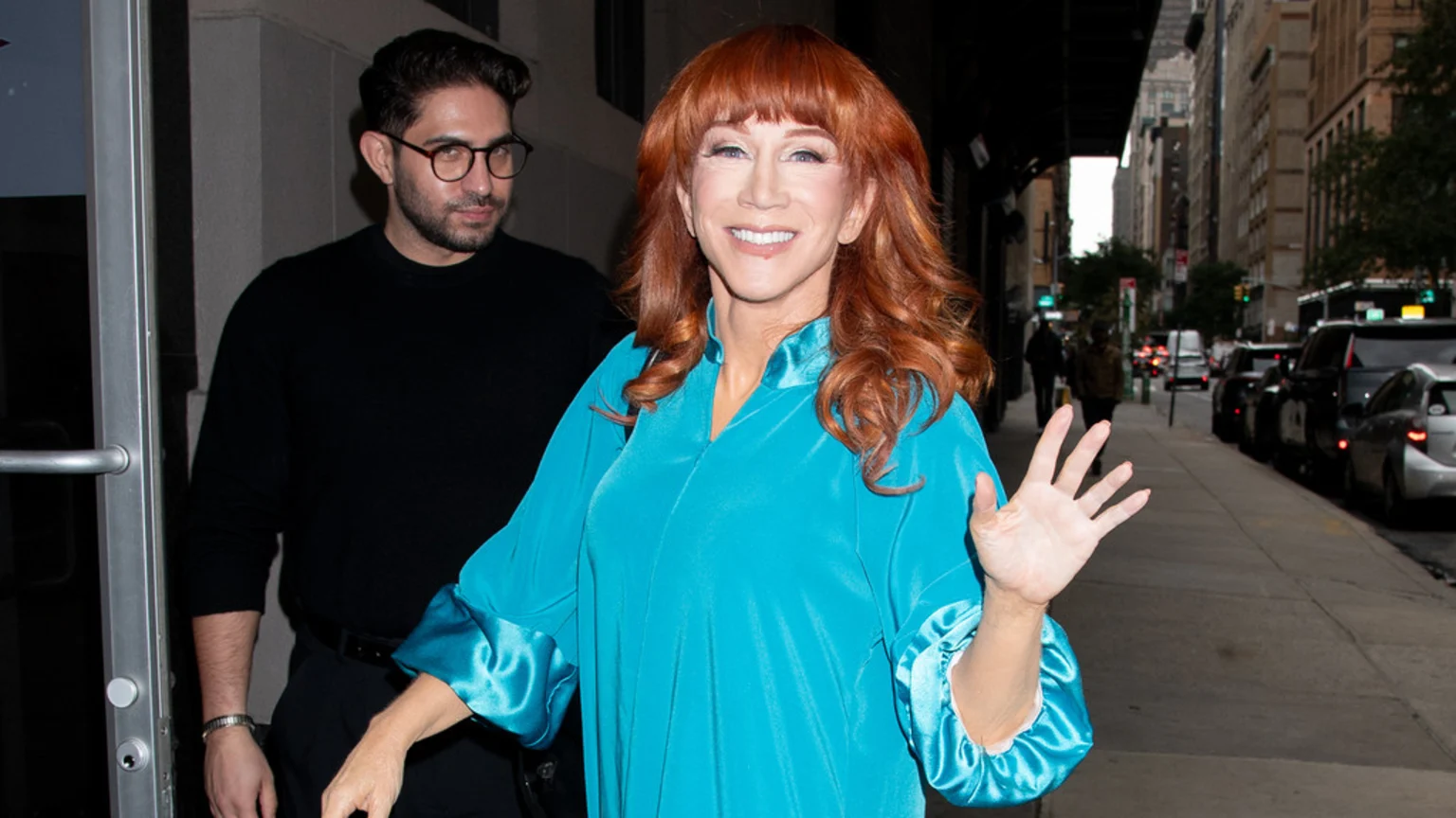 Kathy Griffin with red hair smiles and waves, wearing a bright blue tunic, while a man in glasses and a black sweater stands behind her on a city street.