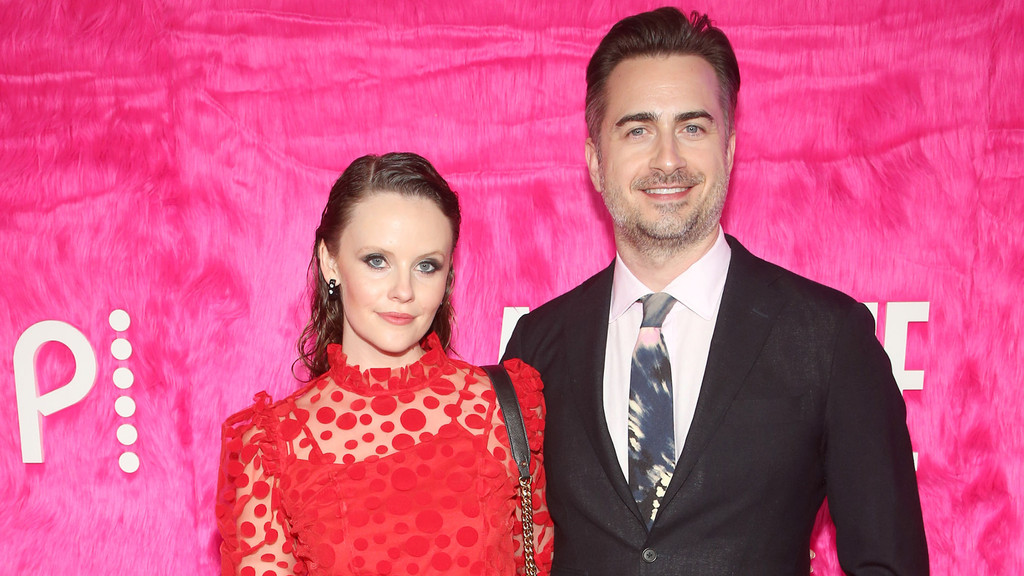 Sarah Ramos in a red polka-dot dress and Matt Spicer in a suit and tie stand smiling in front of a pink furry background.