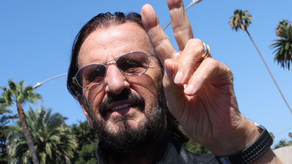 Ringo Starr, wearing sunglasses and a beard, smiles and makes a peace sign against a bright blue sky with palm trees in the background.