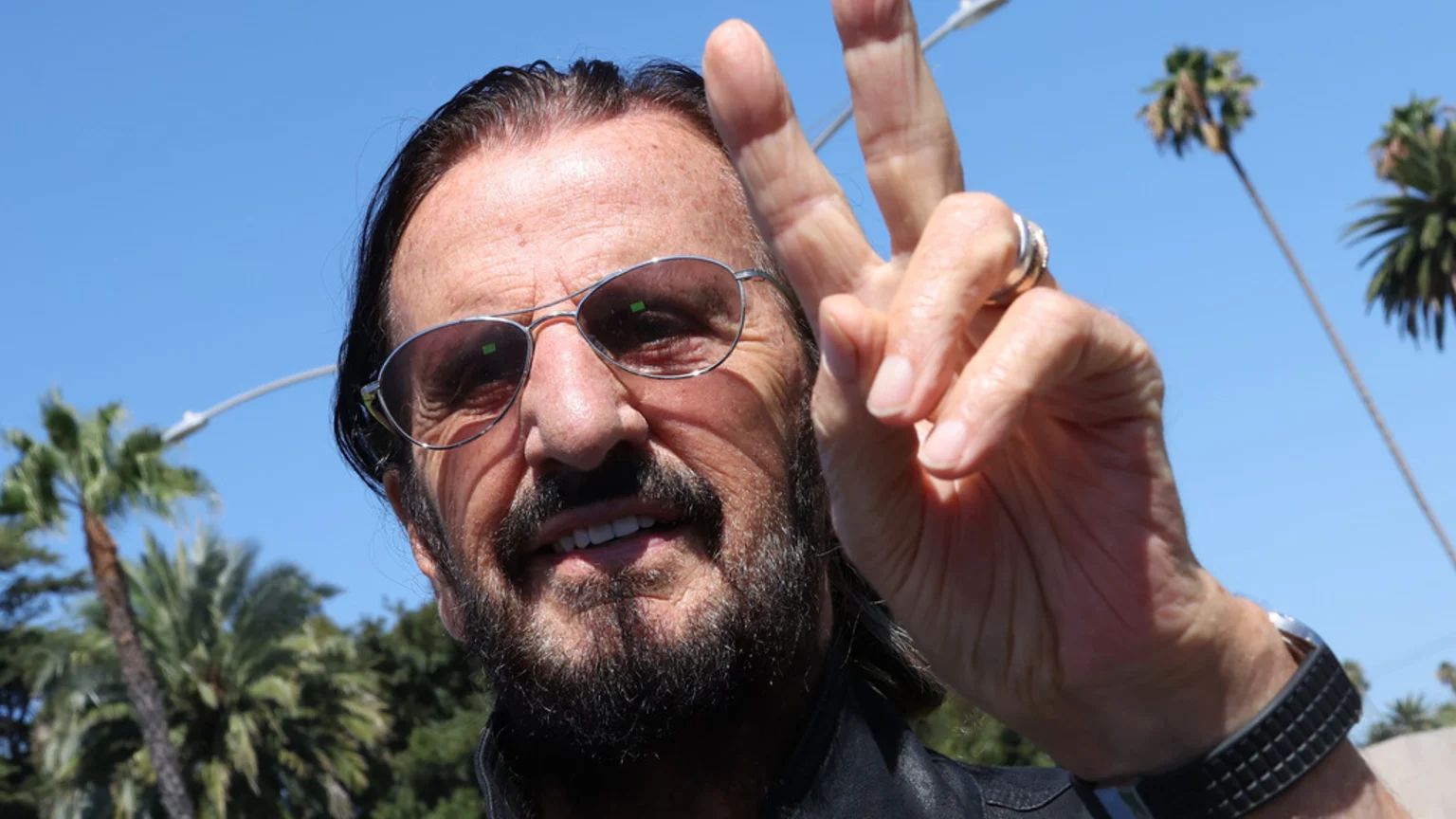 Ringo Starr, wearing sunglasses and a beard, smiles and makes a peace sign against a bright blue sky with palm trees in the background.