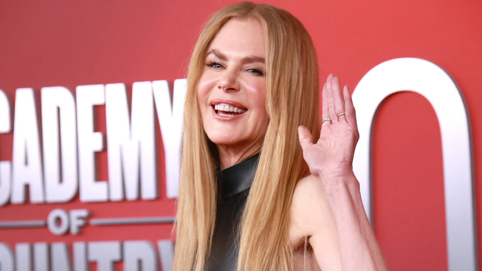 Nicole Kidman, with long blonde hair, smiles broadly with an open mouth and raised hand, against a red backdrop with white letters spelling "ACADEMY OF COUNTRY".