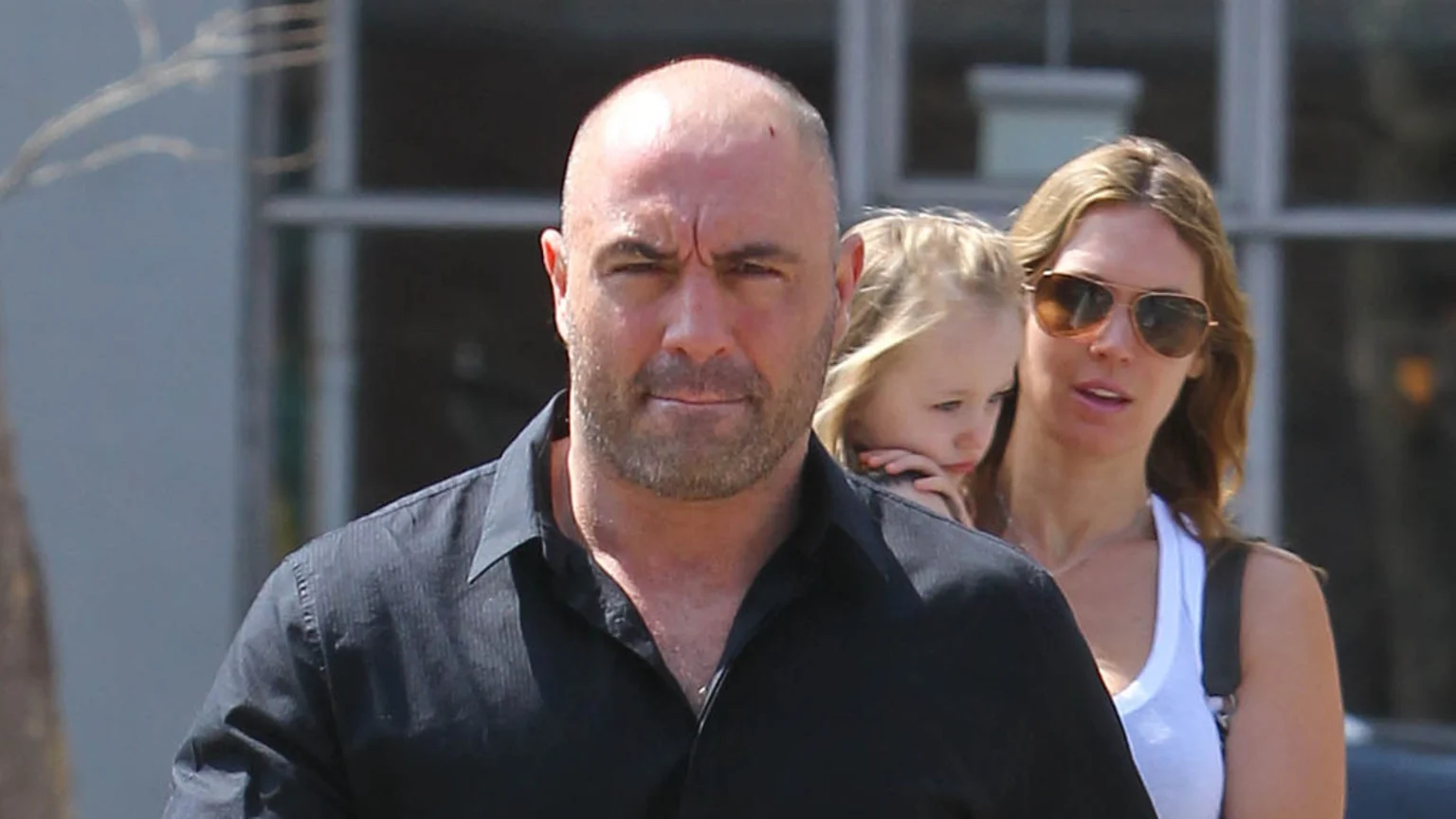 An outdoor candid photo showing Joe Rogan in the foreground wearing a dark button-up shirt and looking at the camera. Behind him, a woman with sunglasses holds a young child in an urban or suburban street scene during bright daylight.