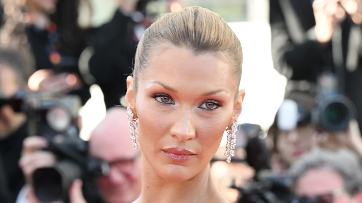 Close-up of Bella Hadid looking forward, wearing prominent makeup and long sparkly earrings, with a blurred background of photographers.