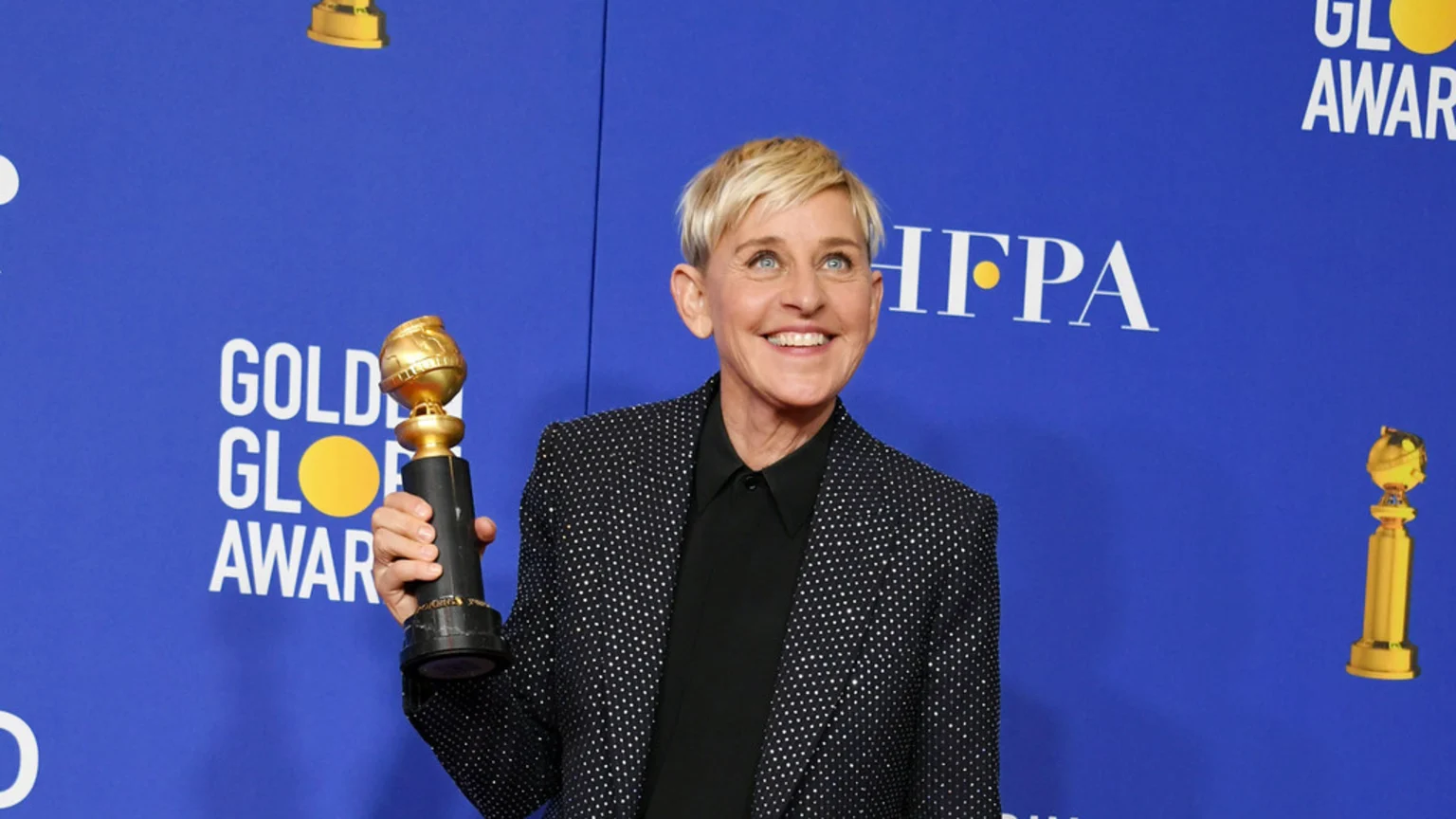 Ellen DeGeneres, with short blonde hair and a sparkly black blazer, smiles brightly while holding a Golden Globe award against a blue backdrop featuring the "Golden Globe Awards" logo.