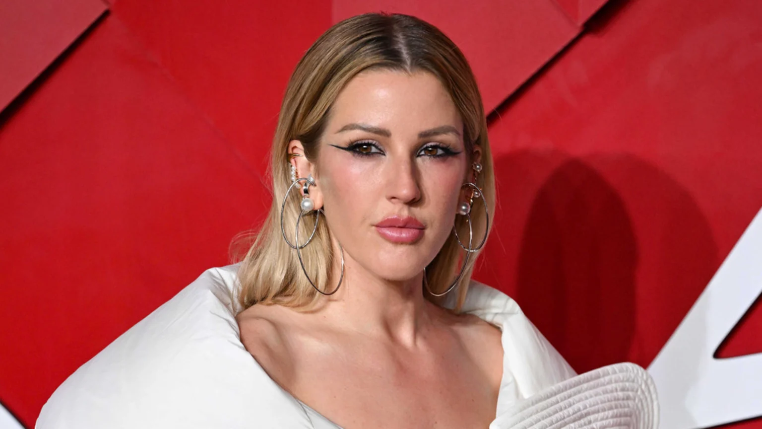 Ellie Goulding with blonde hair and bold winged eyeliner, wearing large hoop earrings with pearls and a white puffy top, against a red background.