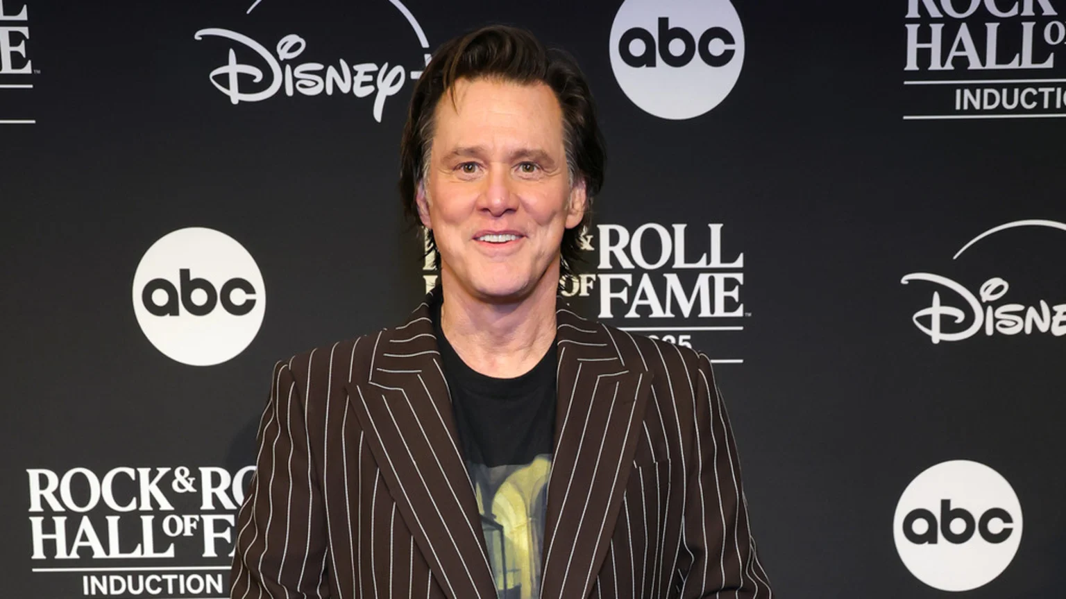 Jim Carrey smiles at the camera, wearing a brown pinstriped suit jacket and a dark t-shirt, standing against a black background with white logos including ABC, Disney, and Rock & Roll Hall of Fame.