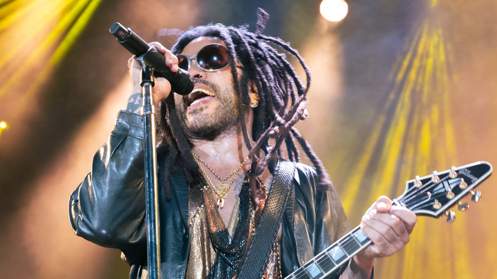Lenny Kravitz performing live on stage, singing into a microphone and holding a Gibson guitar, wearing a leather jacket and sunglasses.