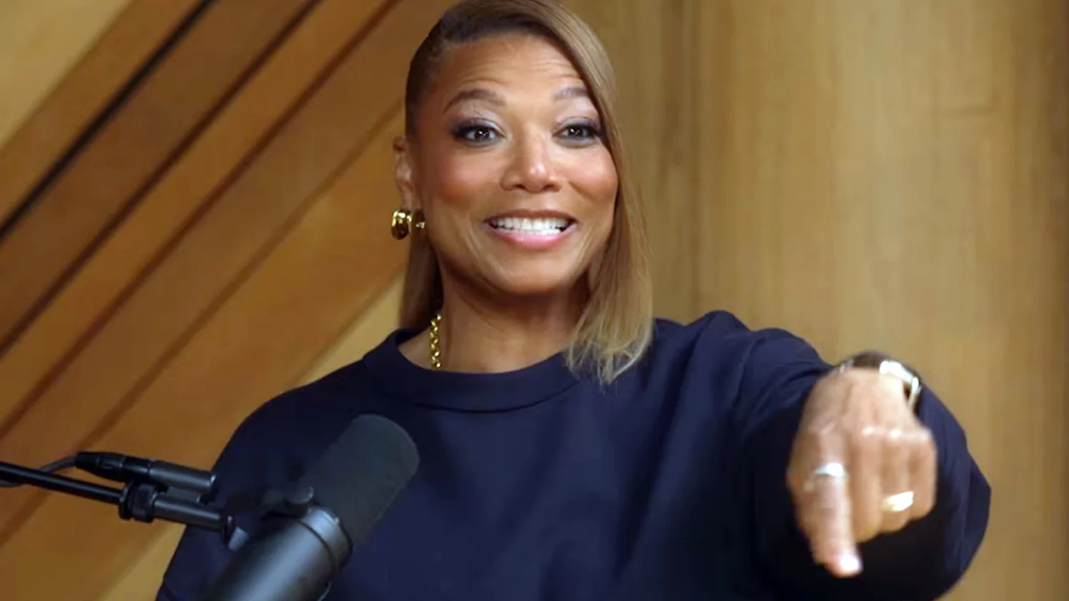 Queen Latifah smiles, wearing a dark blue shirt and gold jewelry, pointing her right hand towards the bottom right while a microphone stands in the foreground.