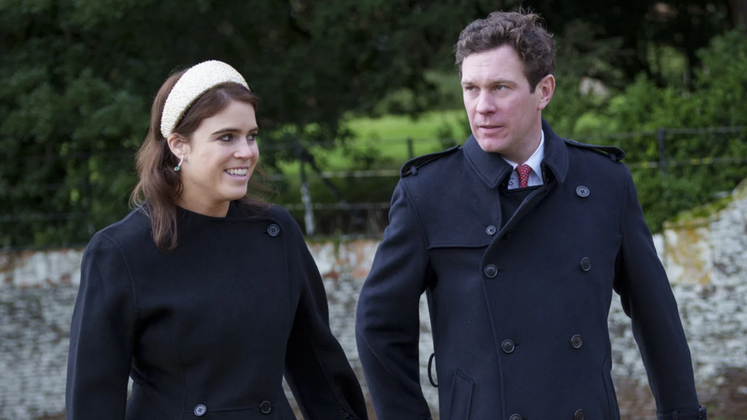 Princess Eugenie, in a dark coat and white headband, smiles next to a man in a dark coat and red tie, walking outdoors with greenery in the background.