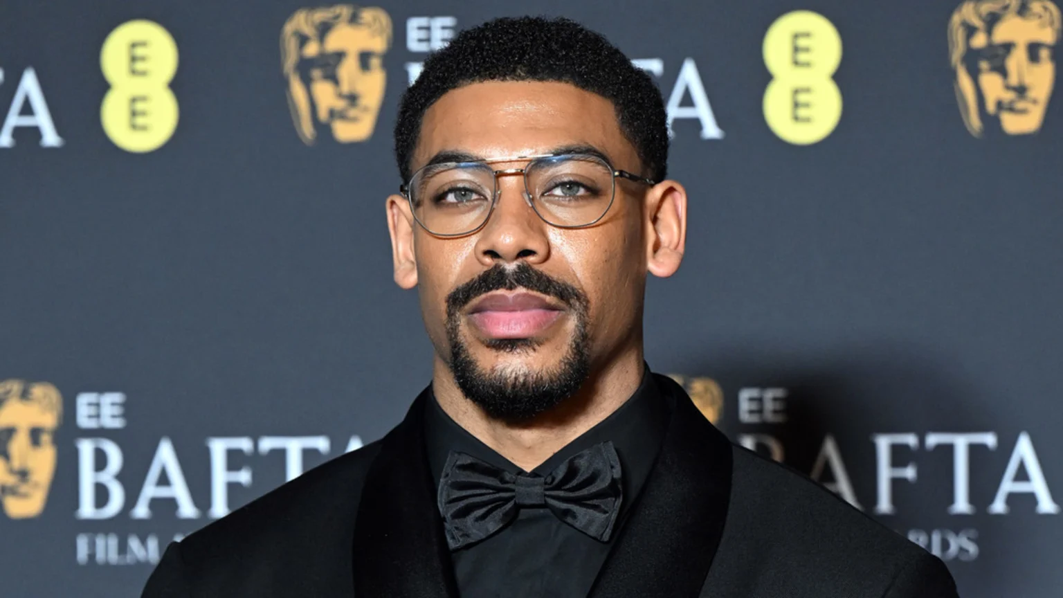 Aaron Pierre, a man with a goatee and glasses, wearing a black suit and bow tie, poses for a headshot in front of a BAFTA backdrop.