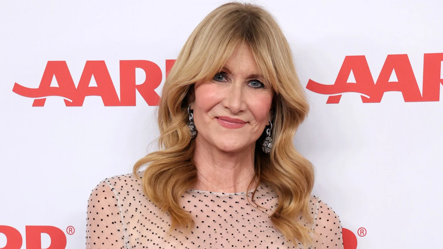 Laura Dern smiles at the camera, wearing a sheer polka dot top and ornate earrings, against a white background with a red AARP logo.