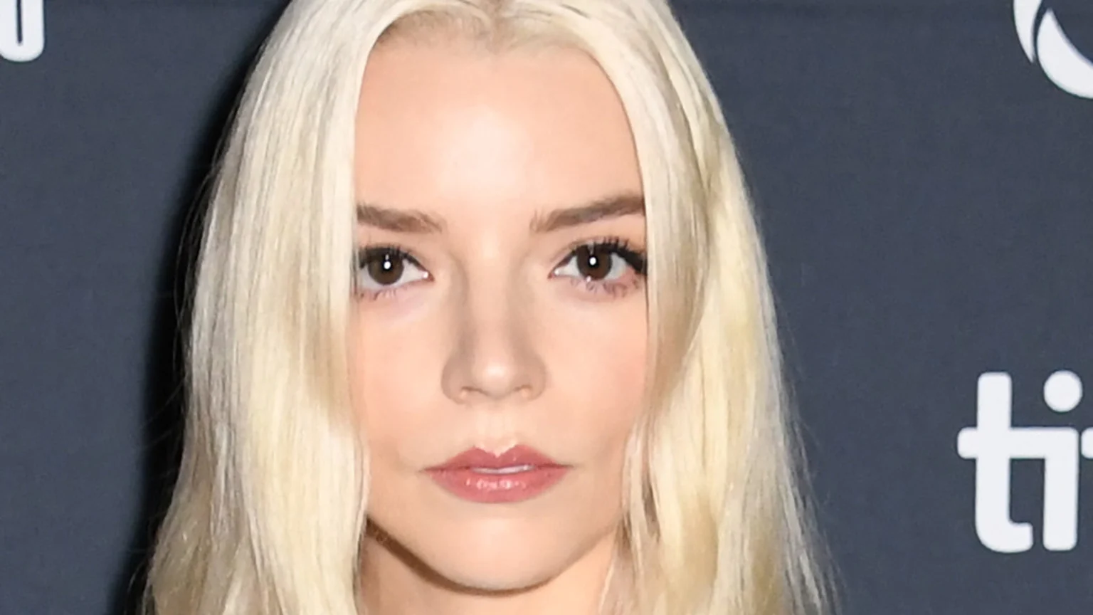Anya Taylor-Joy with long blonde hair, brown eyes, and a neutral expression looks directly at the camera against a dark backdrop with blurred white logos.