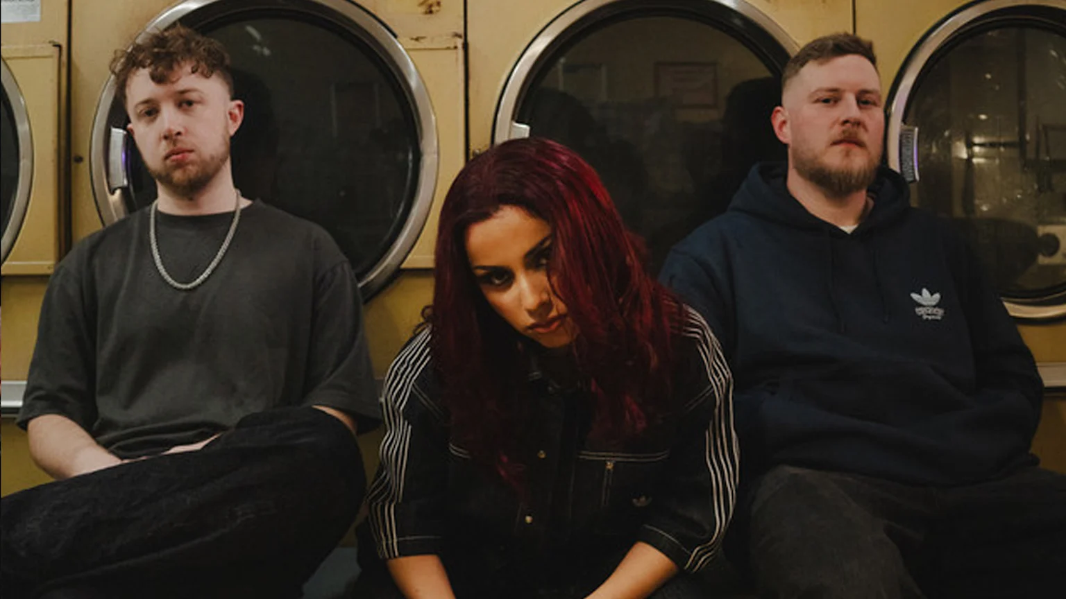 A woman with red hair sits between two men in a laundromat, all looking intently at the camera.