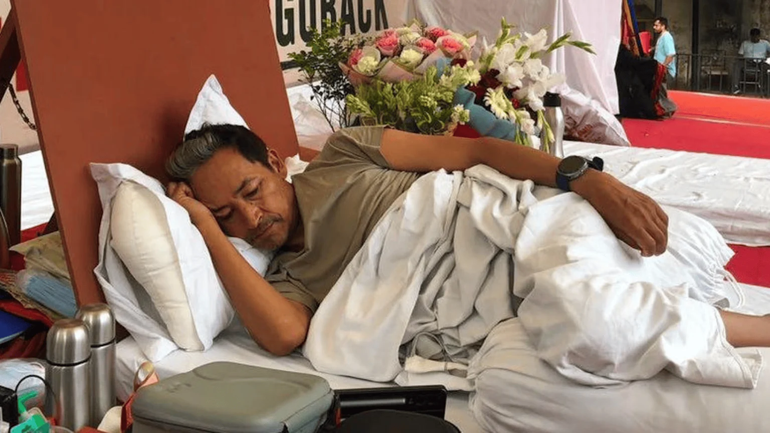 A man lies resting on a bed with white sheets and a pillow, partially covered by a light grey blanket, surrounded by bouquets of flowers, thermos bottles, and personal containers, in a public setting.