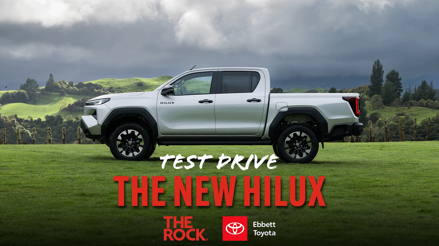 A white Toyota Hilux pickup truck is parked on a green field under a cloudy sky, with "TEST DRIVE THE NEW HILUX" text and "The Rock" and "Ebbett Toyota" logos.