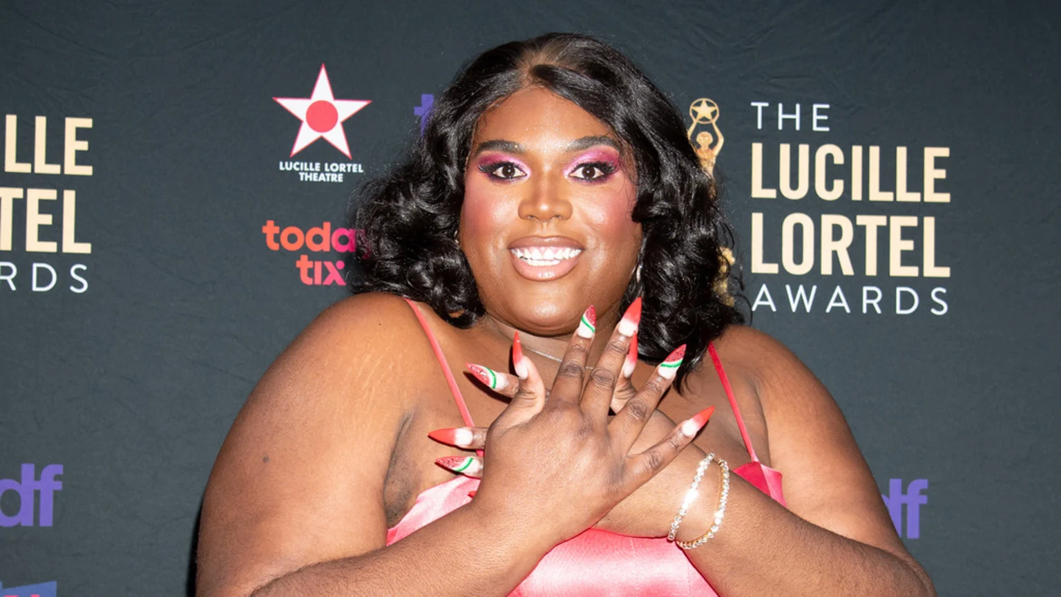A smiling person with dark skin, curly dark hair, and bright pink eye makeup, wearing a red dress and long, watermelon-themed nails, poses in front of a Lucille Lortel Awards banner.