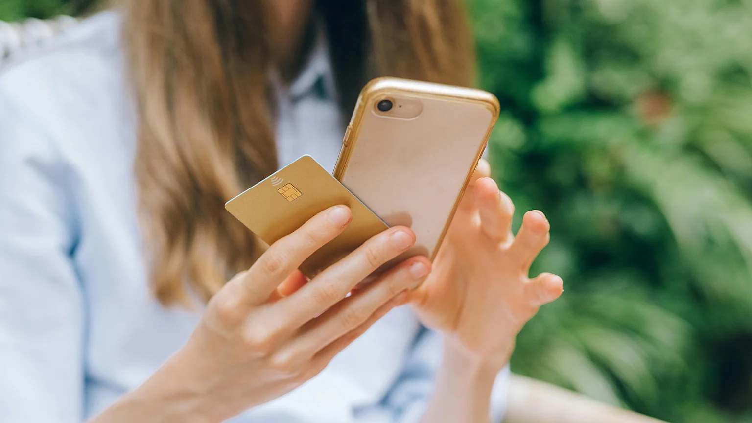 Close-up of hands holding a gold smartphone and a gold credit card with a chip and contactless payment symbol, against a blurred green background.