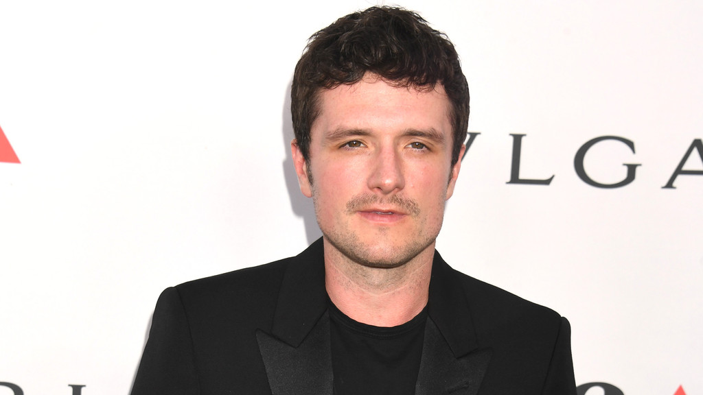 Josh Hutcherson looks forward with short dark curly hair, a light mustache and beard, wearing a black suit jacket and t-shirt, against a white background with a blurred "BVLGARI" logo.